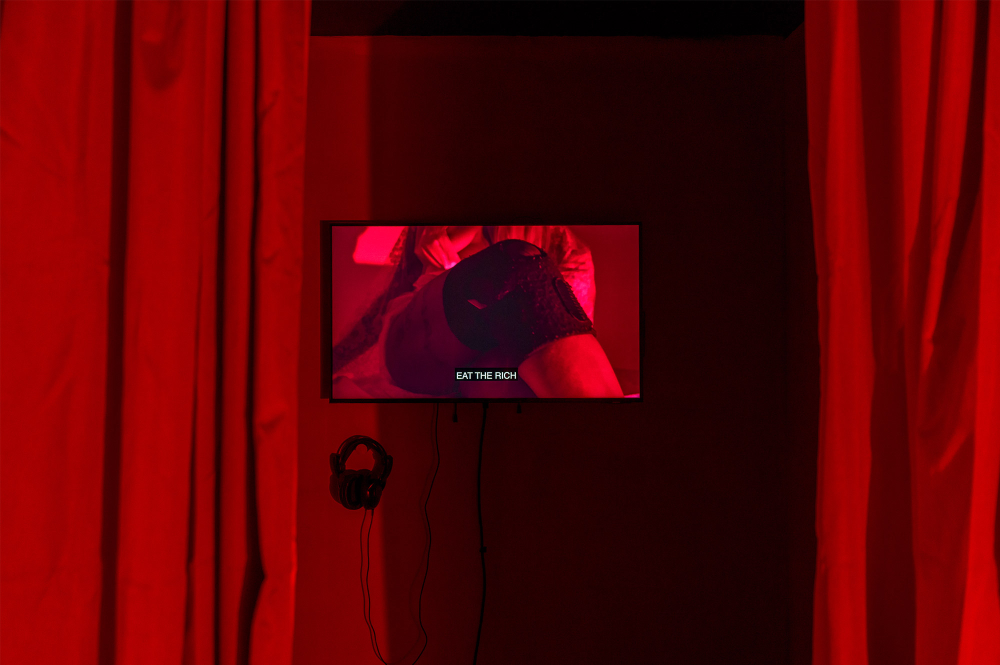 Red curtains surrounding a video installation. A knee is visible, with the text EAT THE RICH