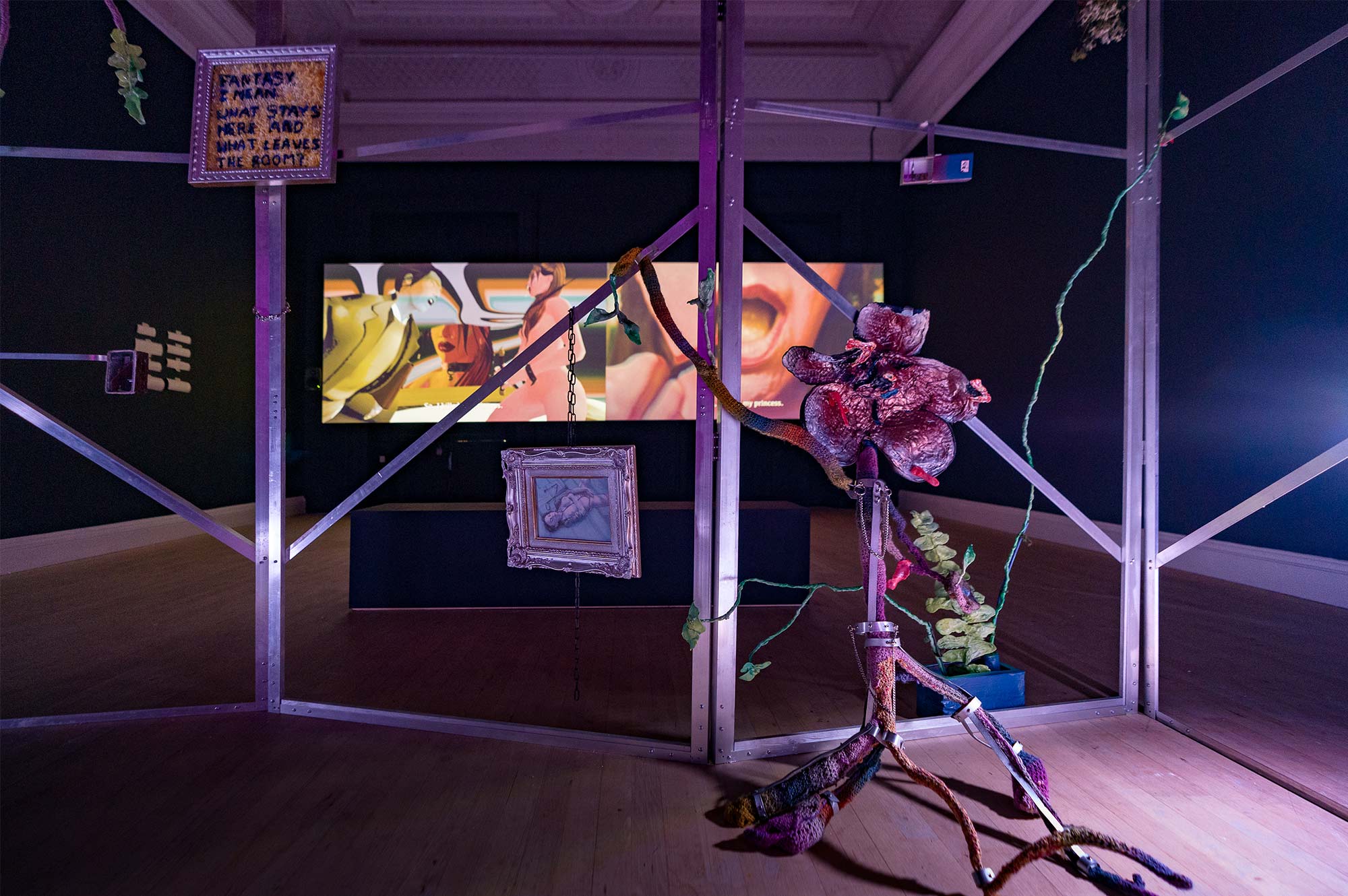 Metal frames surround a mutant flower sculpture. Behind them is a video piece displaying a 3D-rendered sex worker.