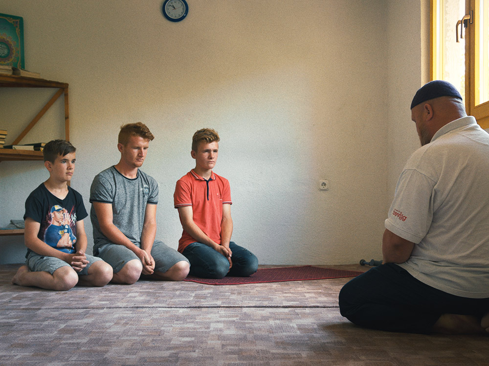 Three white boys kneel in a prayer room in front of their father wearing religious headpiece
