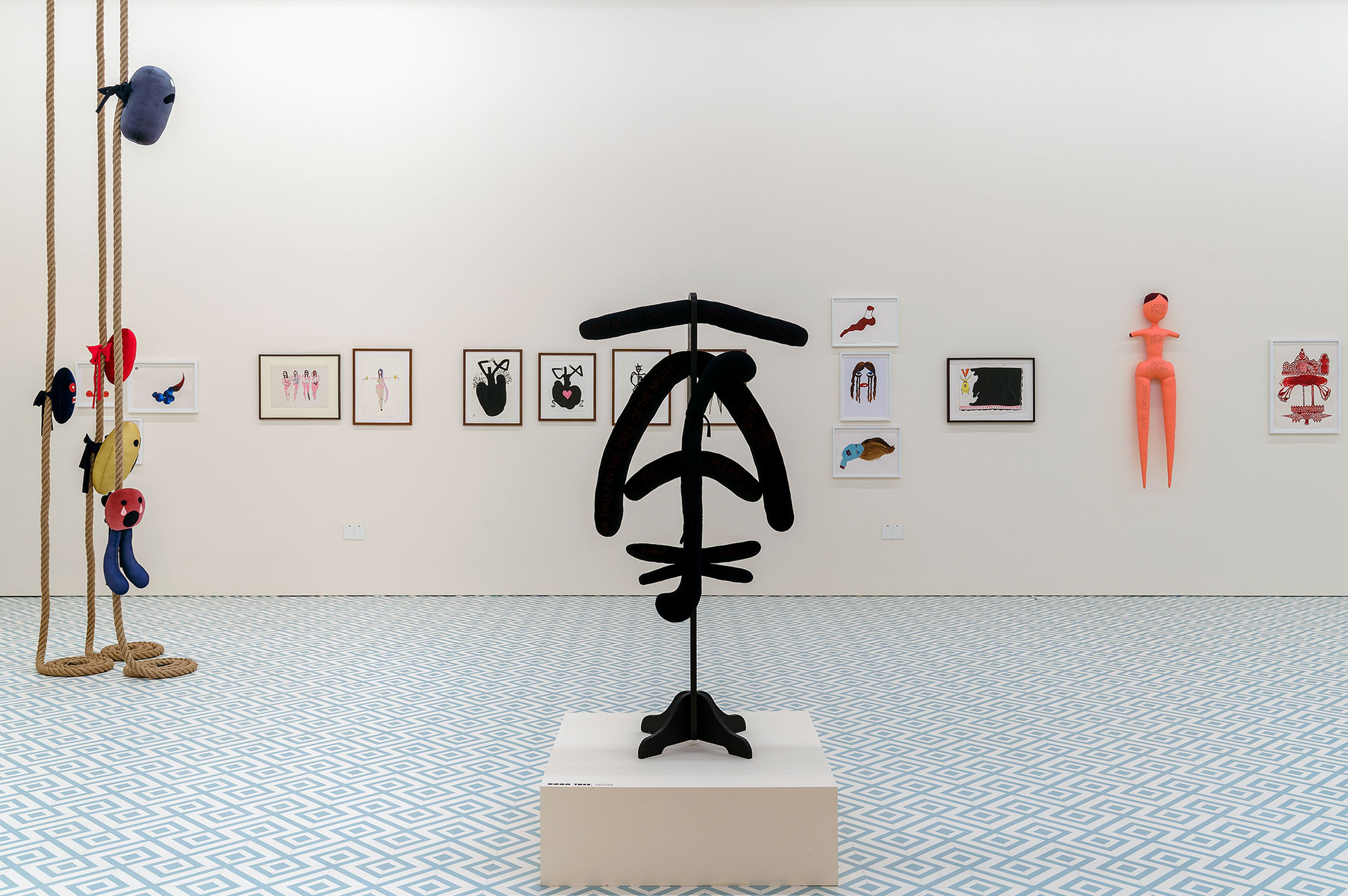 A 'tree' of black fabric limbs on a short plinth. On the left, five felt objects hang from three thick ropes hung from the ceiling.