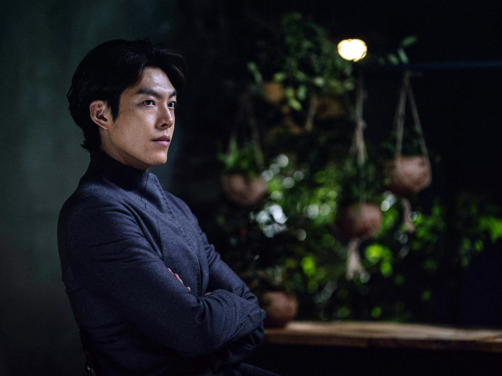 A Korean man in well-tailored clothes looks off-camera, arms crossed, in front of a wall of hanging plants.