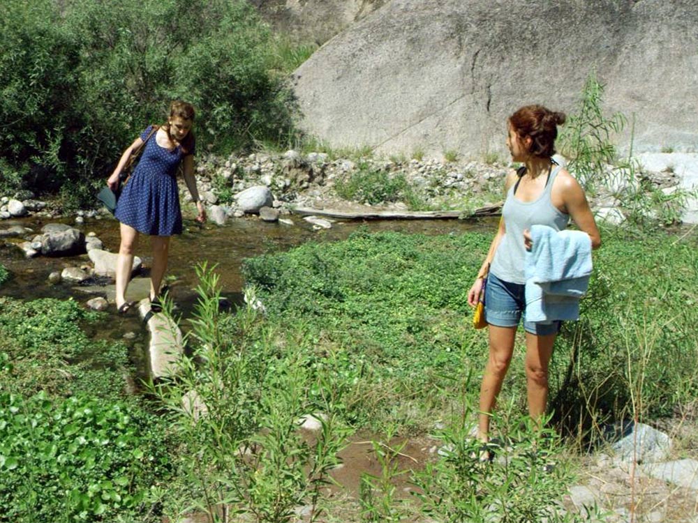 Two women cross a stream surrounded by lush grass