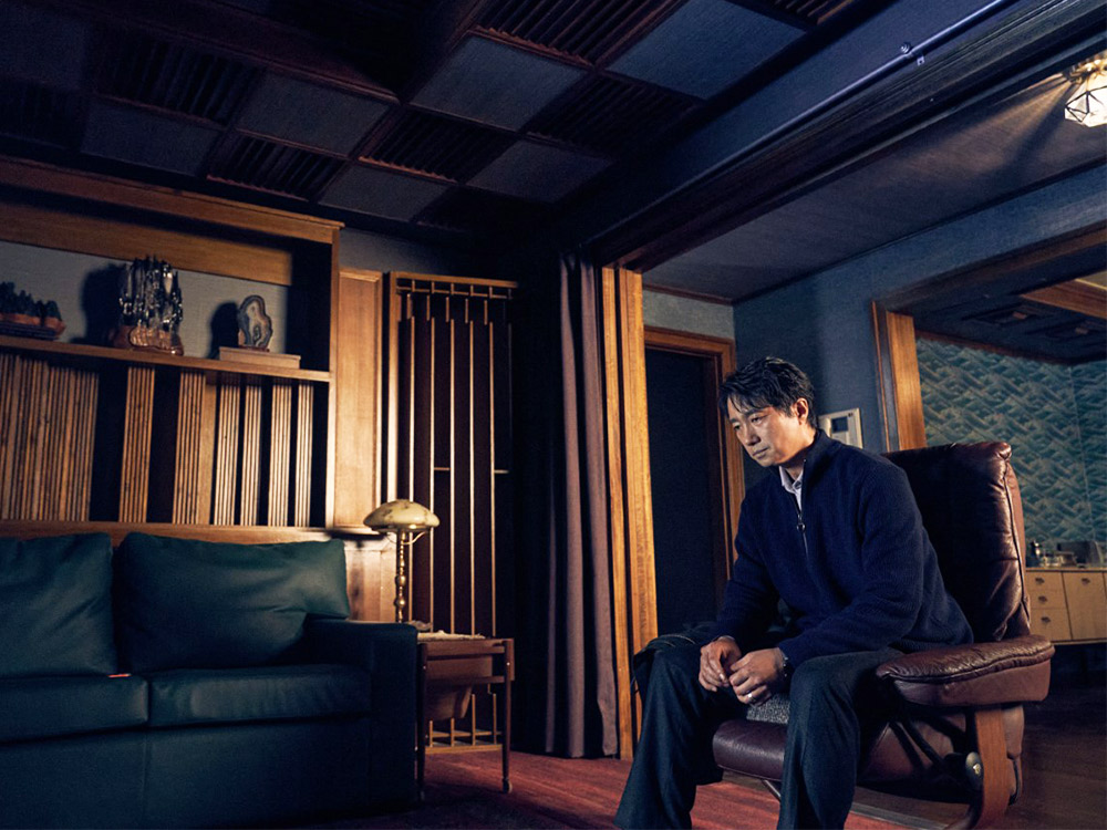 A Korean man sits in a dimly lit living room fitted with antique furniture. He is sitting at a strange angle, staring down into the floorboards