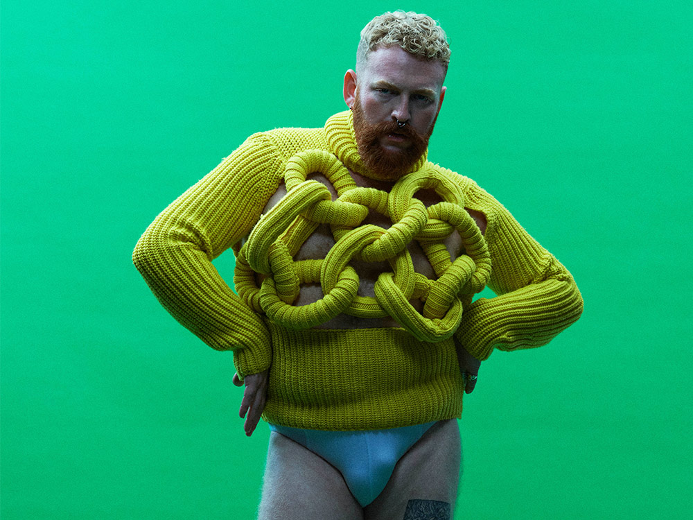 A bearded person with a bright knitted top, wearing blue trunks, against a bright background. They're posed with arms against hips