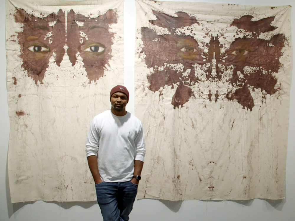 A black man stands against two hanging murals of his work. They look like brown rorschach patterns with eyes.