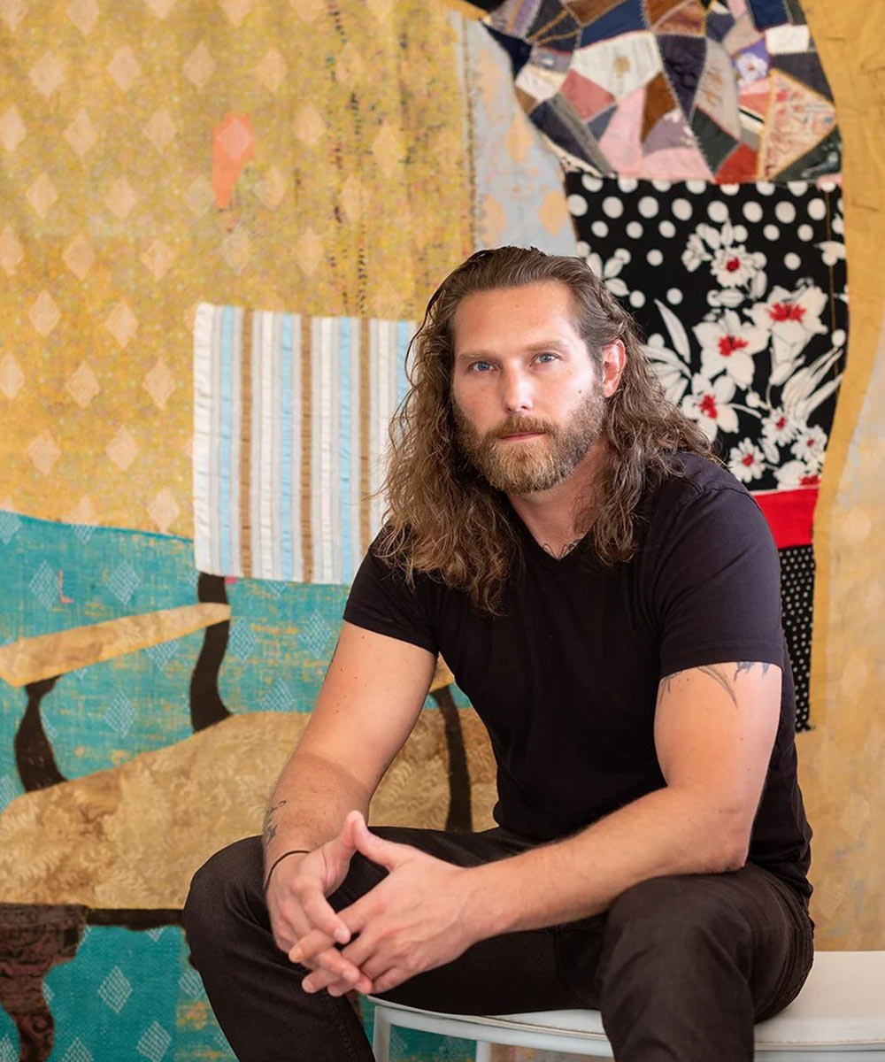 Artist Jesse Krimes, a white man with long curled hair, sits in front of his work and stares into the camera
