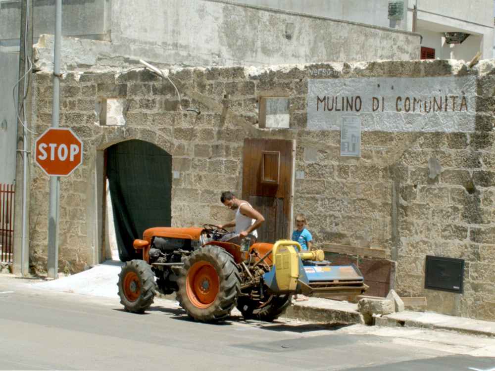 A man in a singlet sits on a tractor in front of a low brick wall. A child is standing by, looking at the tractor
