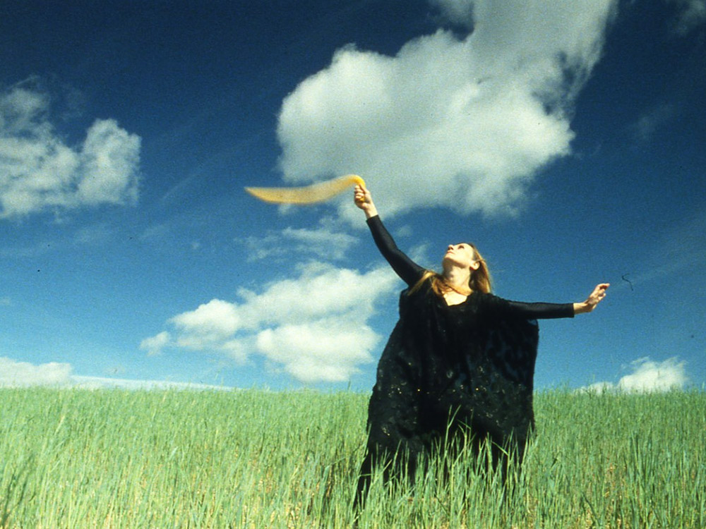 A person with long hair in a flowing black outfit waves a yellow cloud in the air. They are wandering through a grassy field with a blue, clouded sky