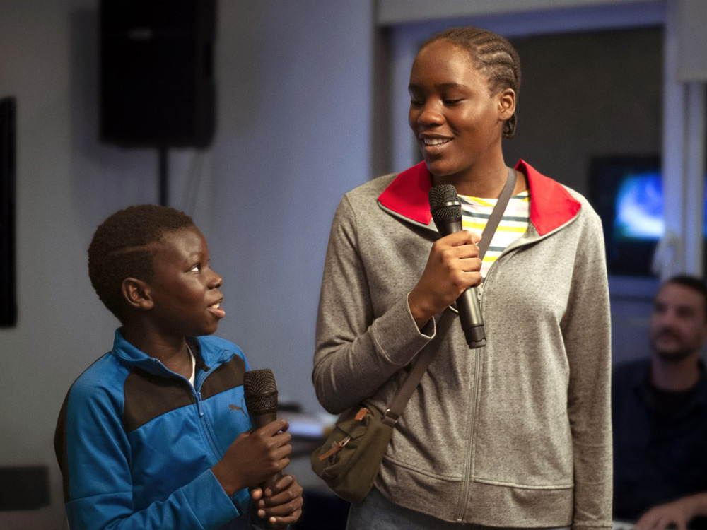 A brother and sister dressed in casual clothes stare towards eachother encouragingly, holding microphones and presenting to an audience