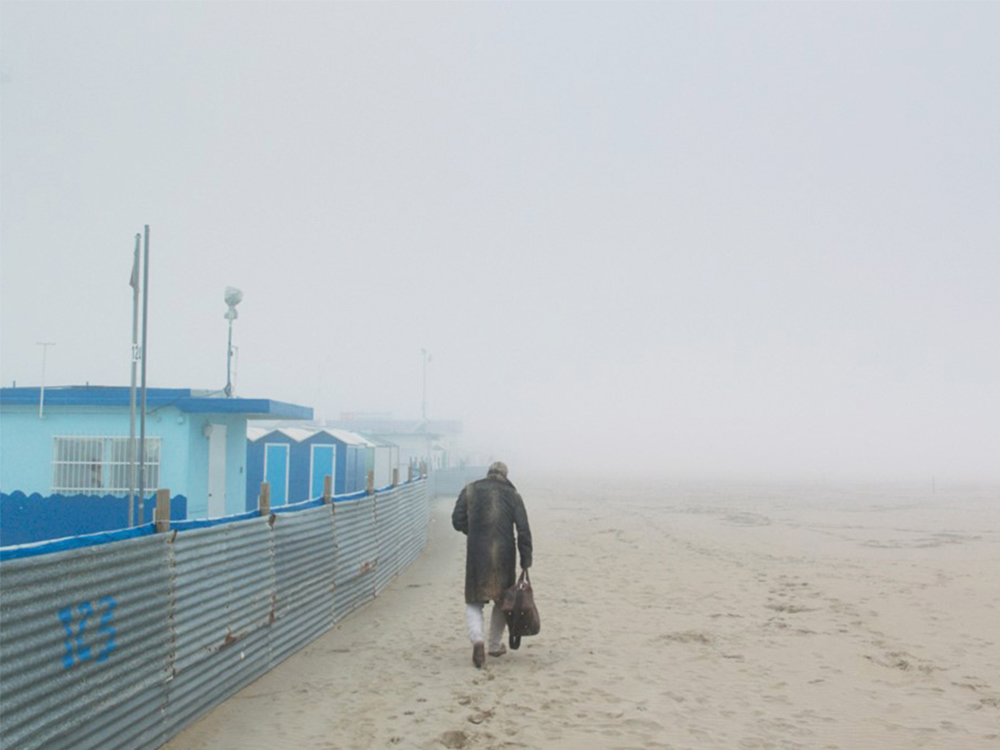An old man in a coat carries a duffle bag. He's walking into the fog along a quiet beach, with a few blue cabins by the sand