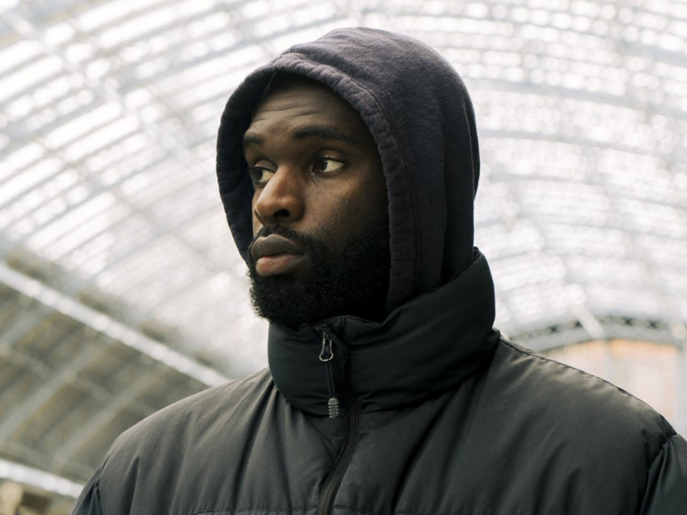 A headshot of Bawo, wearing a winter jacket and hoodie, against the hatched ceiling of Kings Cross Station