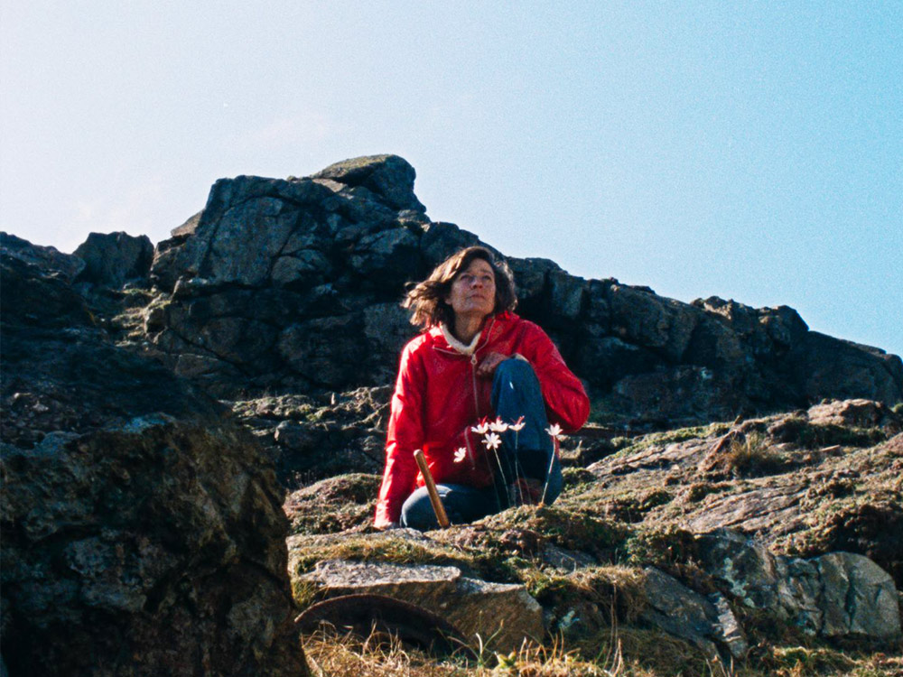 A person in a wind jacket and jeans sits atop a mossy rock. The image is shot on grainy colour film.