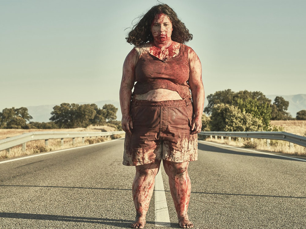 A person in bloodstained clothes, blood on their face, stands in the centre of a barren road