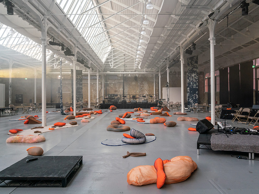 Amorphous orange and brown cushions are spread out across a factory warehouse
