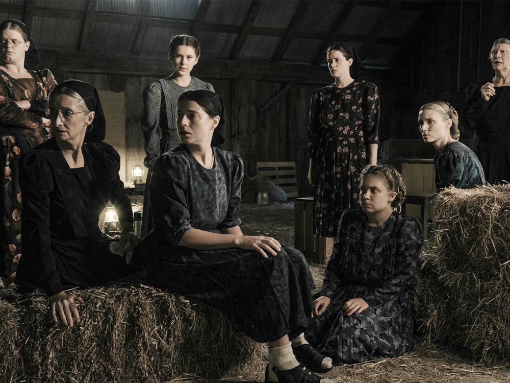 A group of women in a community sit amongst wooden crates and haystacks, looking offscreen in worry. A single person stands staring at the camera, a solid face.