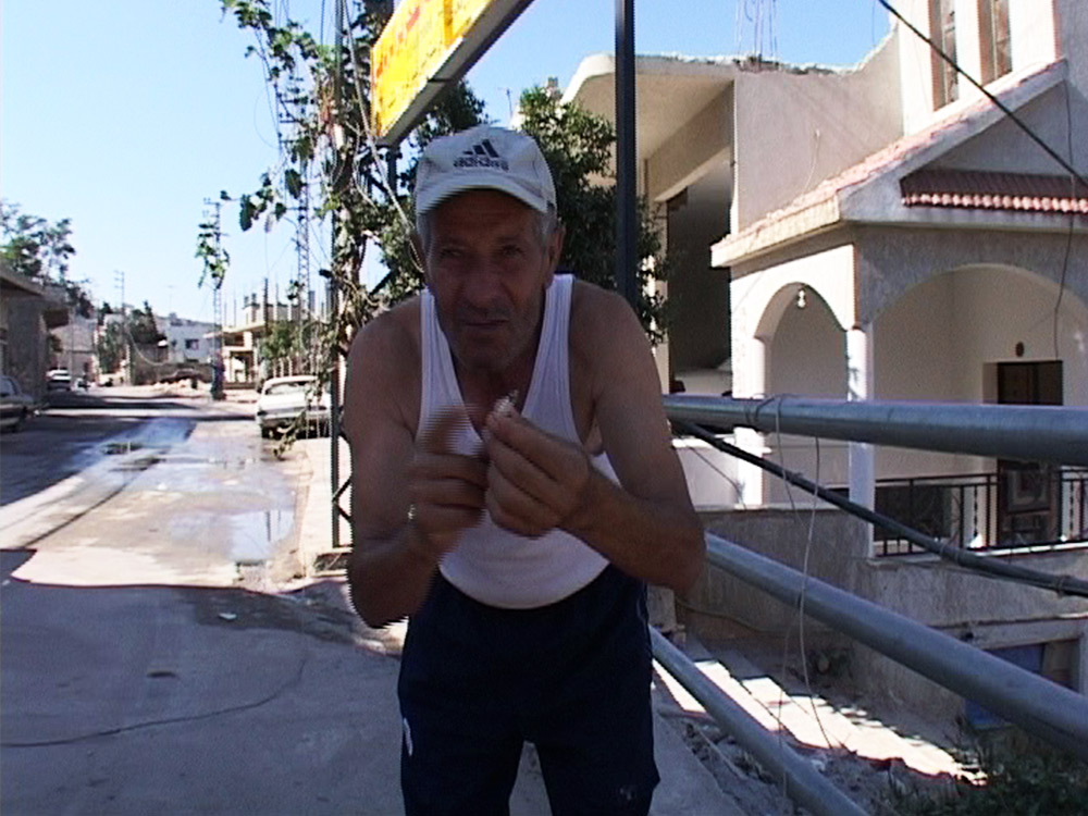 A Lebanese man in a singlet, shorts and adidas cap gestures to something for the handheld camera. Behind him is the streets of Beirut during the siege in 2006