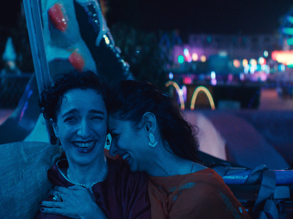 In the night, two people joyfully lean on one another in an amusement park ride