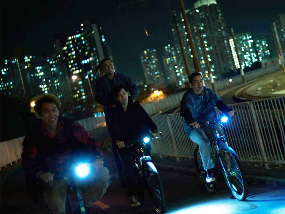 Four people on bikes ride down the road against the bright HK skyscraper skyline
