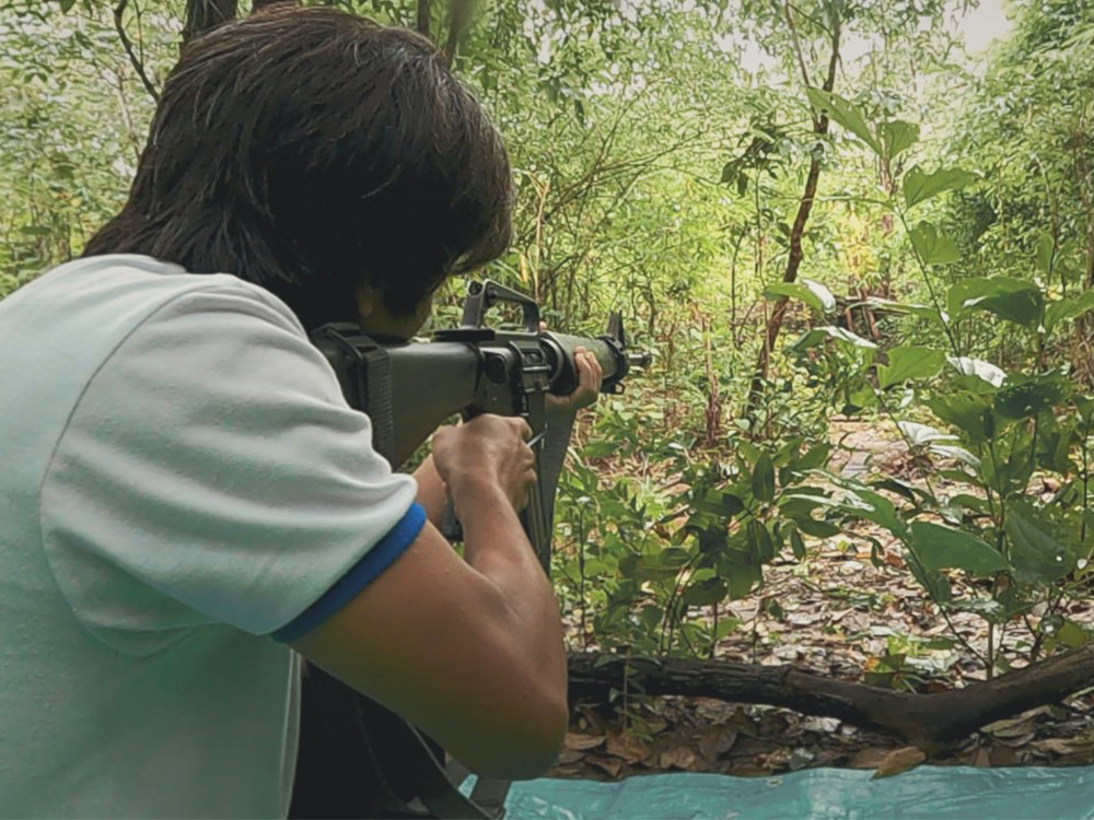 A person in a tshirt aims an assault rifle into the distance in the Burmese jungle