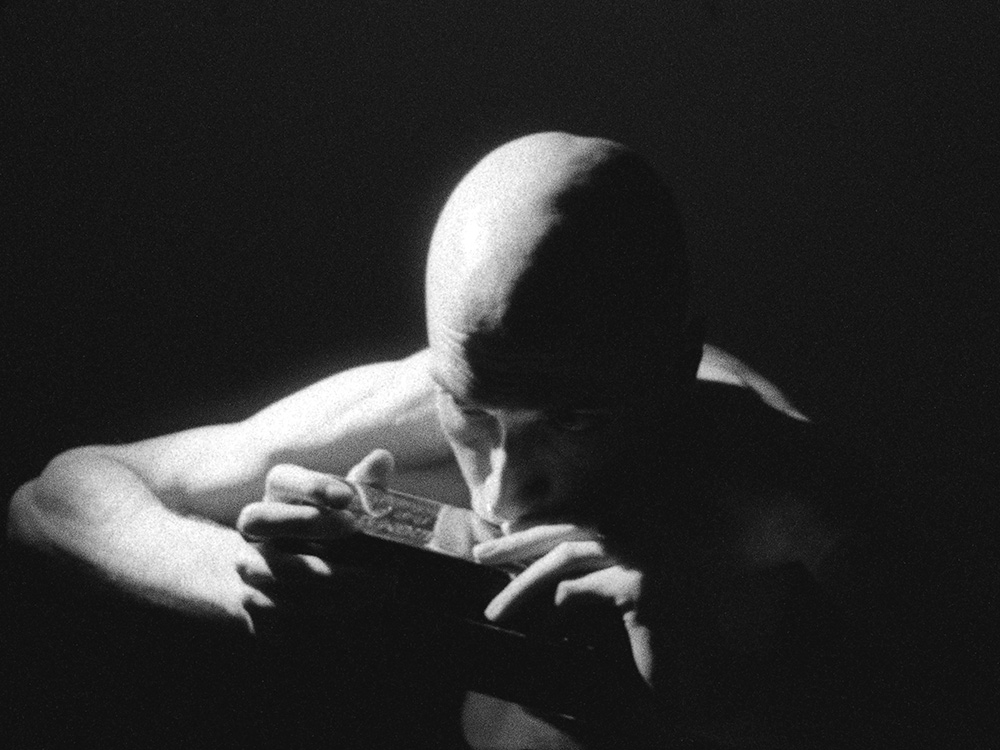 Daniel Blumberg blows into a harmonica, shirtless, in the dark