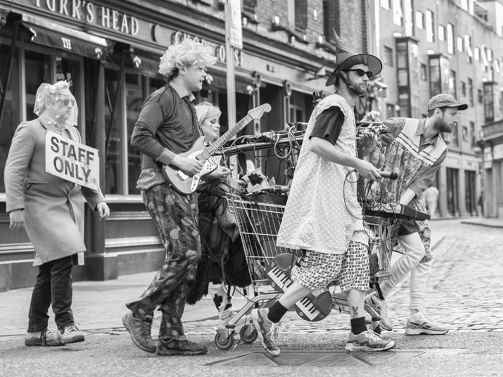 The band Acid Granny dressed in scattered clothes, with instruments and electronic gear dangling from a shopping trolley through the streets