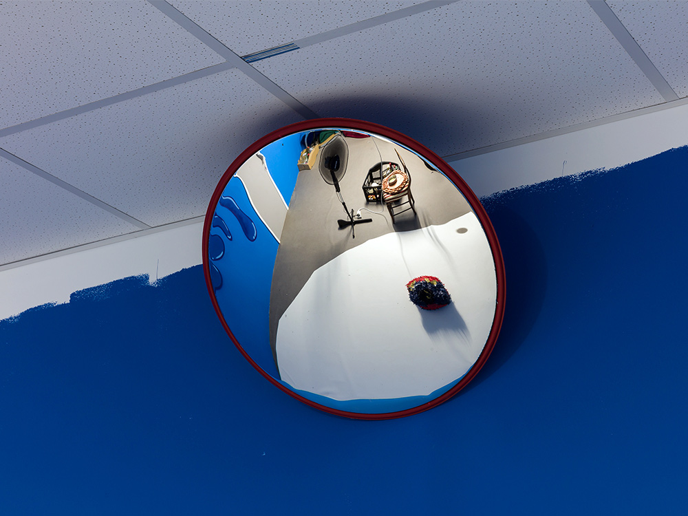 A round mirror reflecting a blue room from the exhibition, showing a fan and a stool