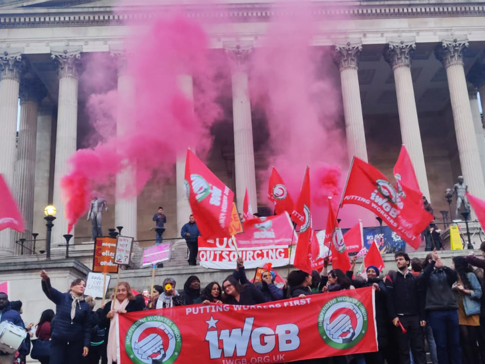 A large crowd of IWGB union members fly red flags and let off pink smoke in front of a building, protesting