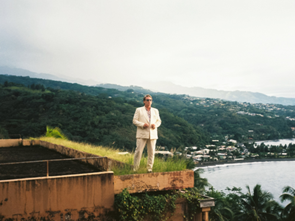 A man in a white suit and sunglasses stands on a rooftop overlooking a pink island sunset