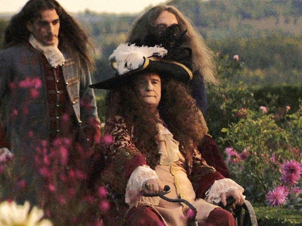 The ageing King Louis XIV sits on a chair flanked by his servants in a lush garden full of pink sunburst-shaped flowers
