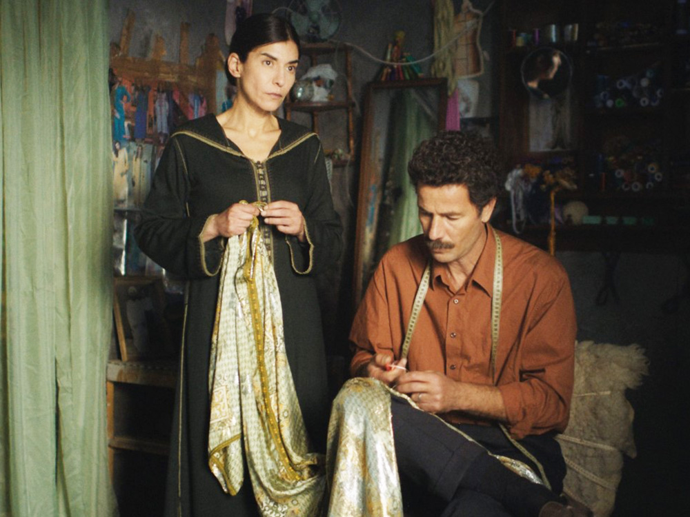 A man and woman couple work on pieces of fabric in their workshop/home filled with shelves of materials