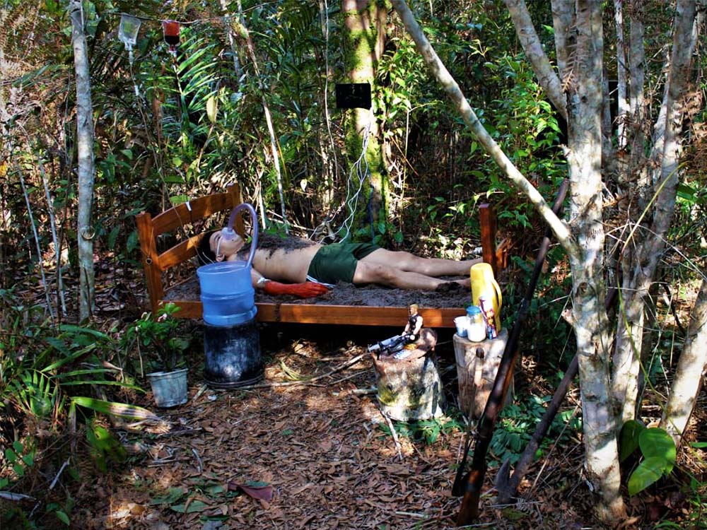 A person with dirt on his chest rests in a bed covered with dirt, in a jungle. He has a pipe attached to his mouth, which is receiving water from a small water cooler tank.
