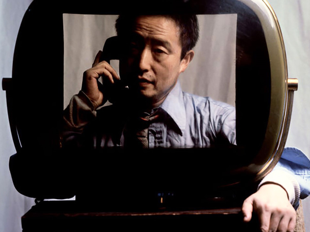 Nam June Paik, a Korean man, holds a phone receiver while staring through the screen of an old TV with rounded edges