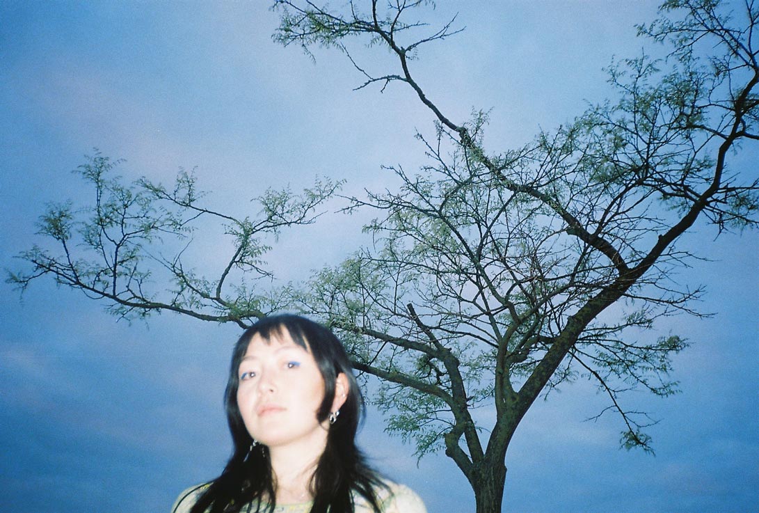 Ans M standing in front of a spindly tree on a grey-blue sky