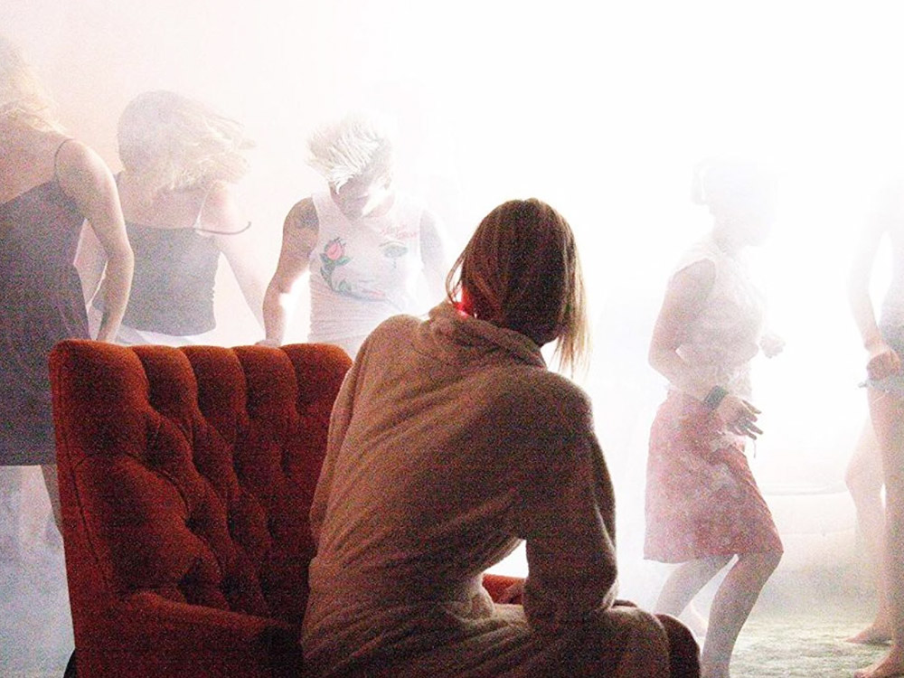 An actress sits on a red chair, back to the camera. She's watching people dance in a bright white light