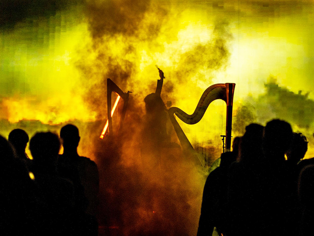 Kety Fusco performs with two harps on stage, surrounded by darkness and yellow fog