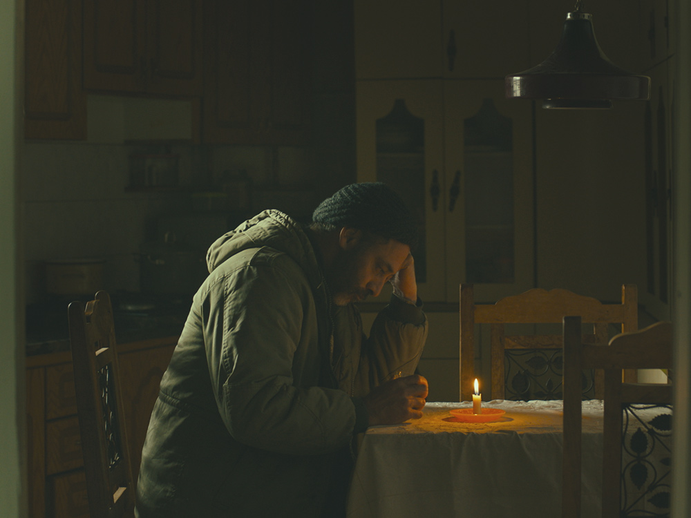 A person in a green coat sits at their kitchen table, writing something by candlelight