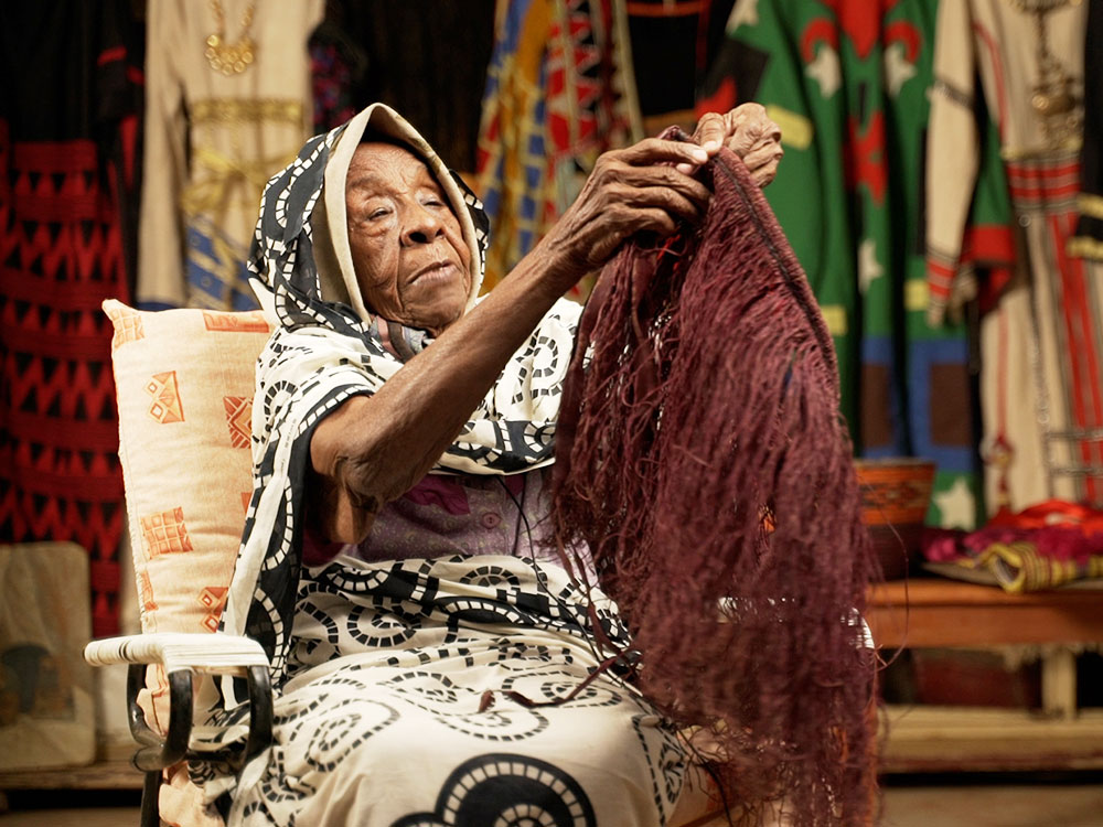 An older woman sits in a chair in front of colourful hanging clothing. She is holding a piece of clothing made of red threads
