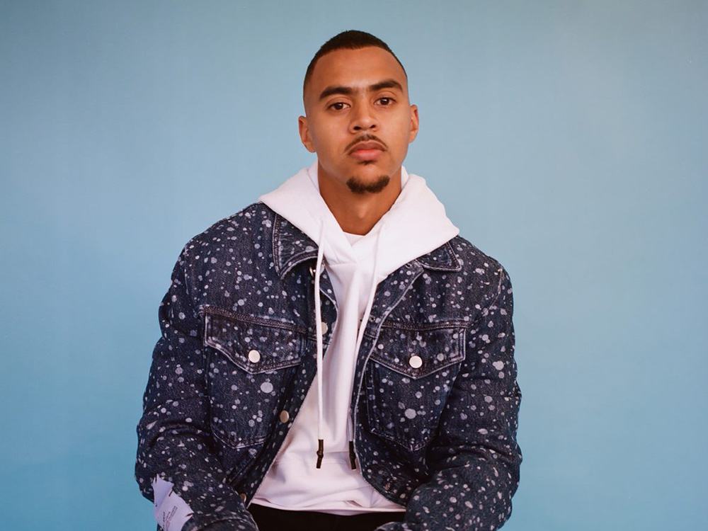 Photo of Swindle, a black man, wearing a white hoodie under a denim jacket, sitting against a blue photo background