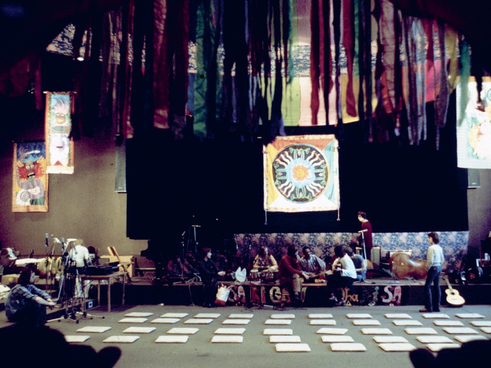 A room covered with Moki's tapestries from the ceiling and on the wall. Cushions are laid out in front of a stage of instruments and mics, ready to perform