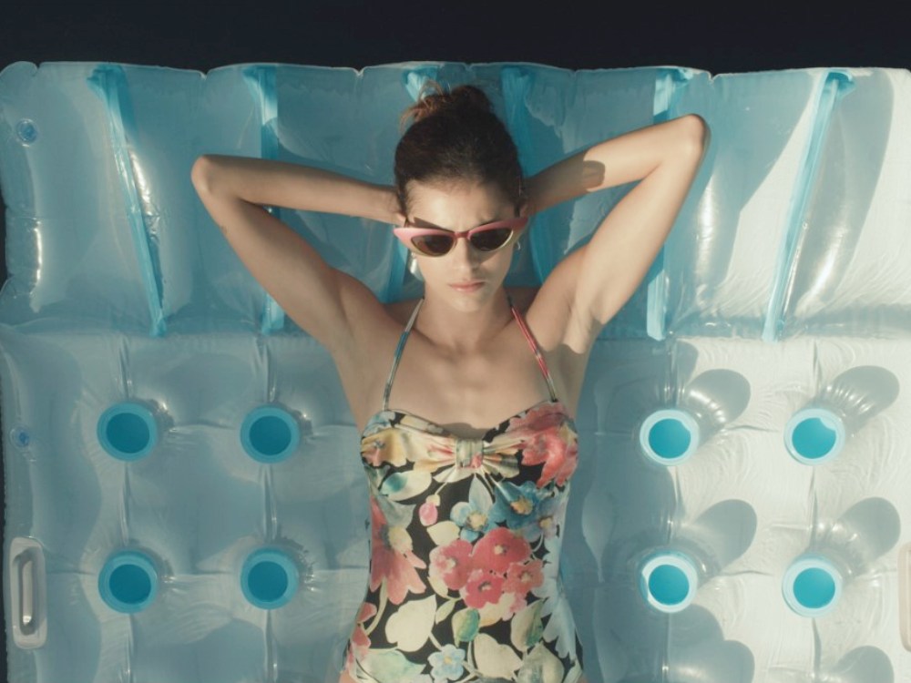 A young twentysomething white woman in sunglasses and bathing suit lies on a blow up inflatable bed