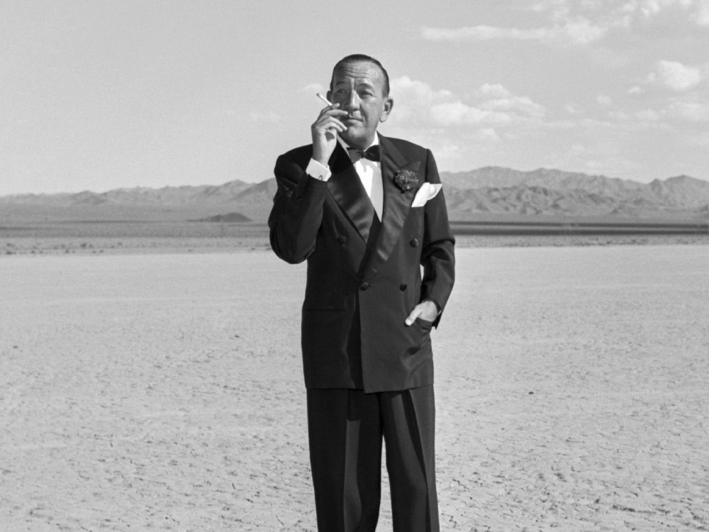 A man in a suit stands on a beach, smoking a cigarette, hand in pocket