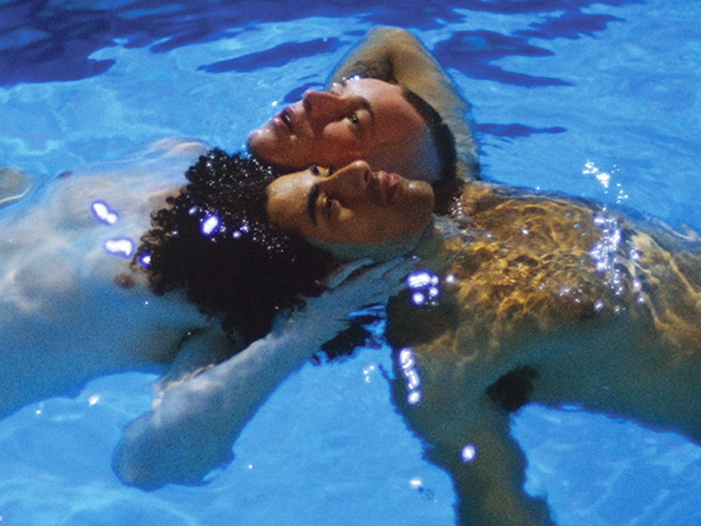 Two young boys float in the pool, their heads up, interlocked, under bright sunlight