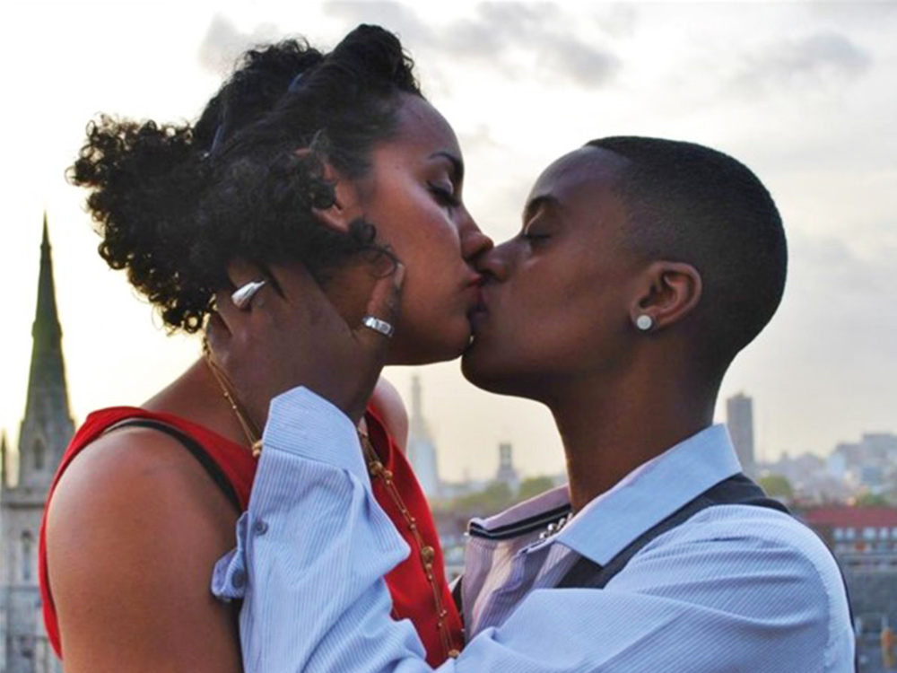 Two people kiss against a London sunset of tall concrete buildings and spires