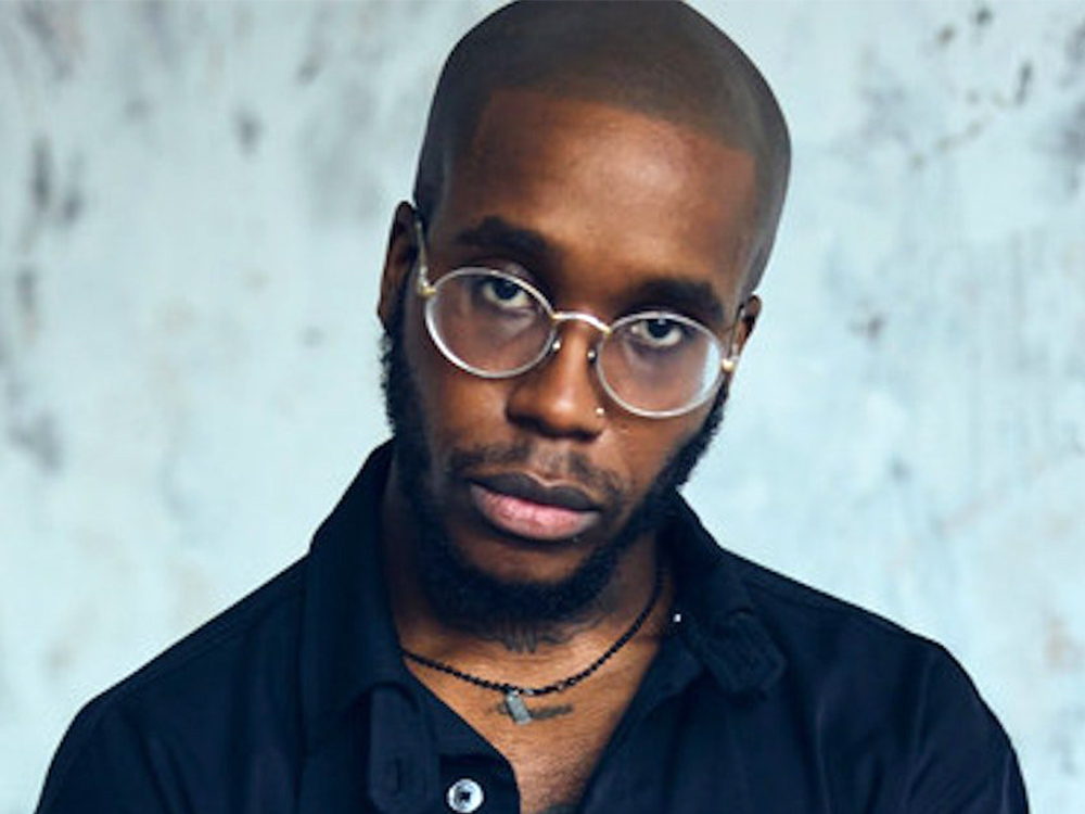 Portrait of Che Lingo. He is Black, wears round glasses, an open navy shirt with a tattoo visible on his chest