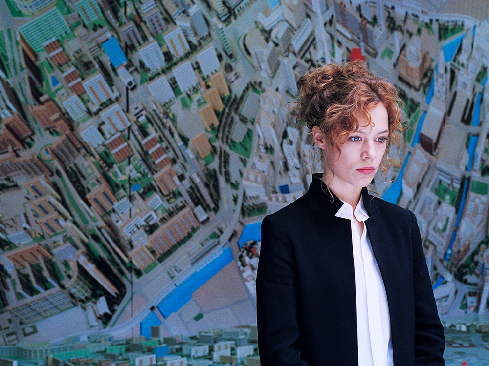 A woman with orange hair and in formal dress stares. Behind her is a top-down map of urban Berlin