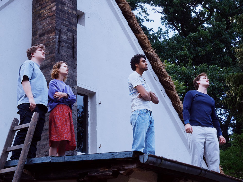 A group of young people stands on the roof of their house, looking off at an unknown disaster
