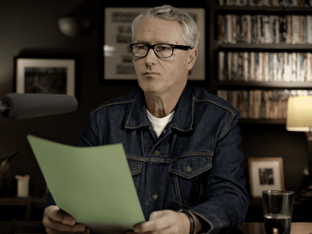 At his desk, a man with grey hair considers a green letter. He's looking away from it