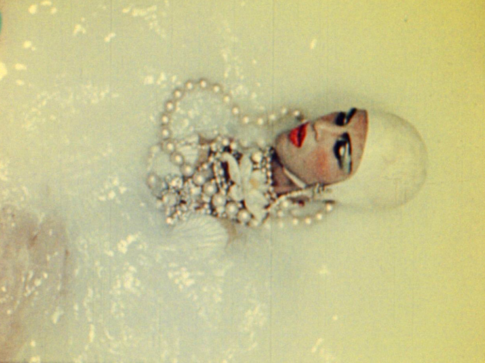 A person with red lipstick, huge pearl necklace and white bodysuit bathes in an opaque white liquid