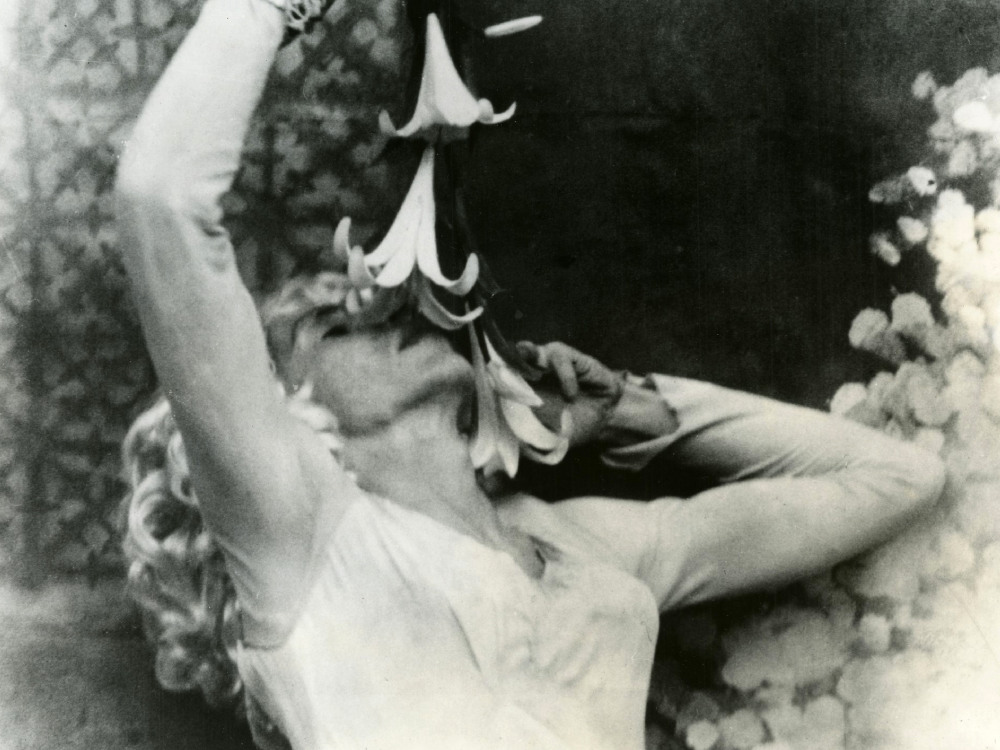 In black and white, a person with long blonde curly hair holds a flower down towards their nose