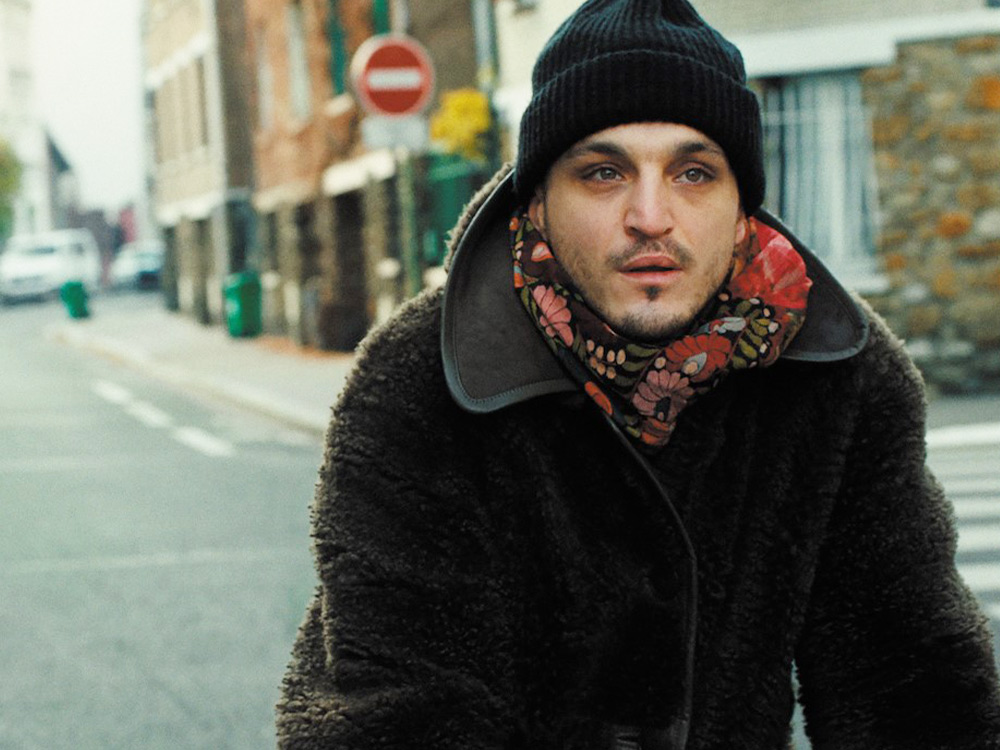 A man in a winter coat, beanie and scarf rides a bike down a backstreet